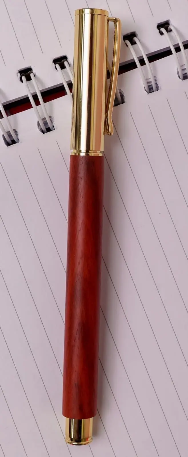 Wooden pen with a reddish-brown barrel, gold cap and clip lying on a spiral notebook, Hensa