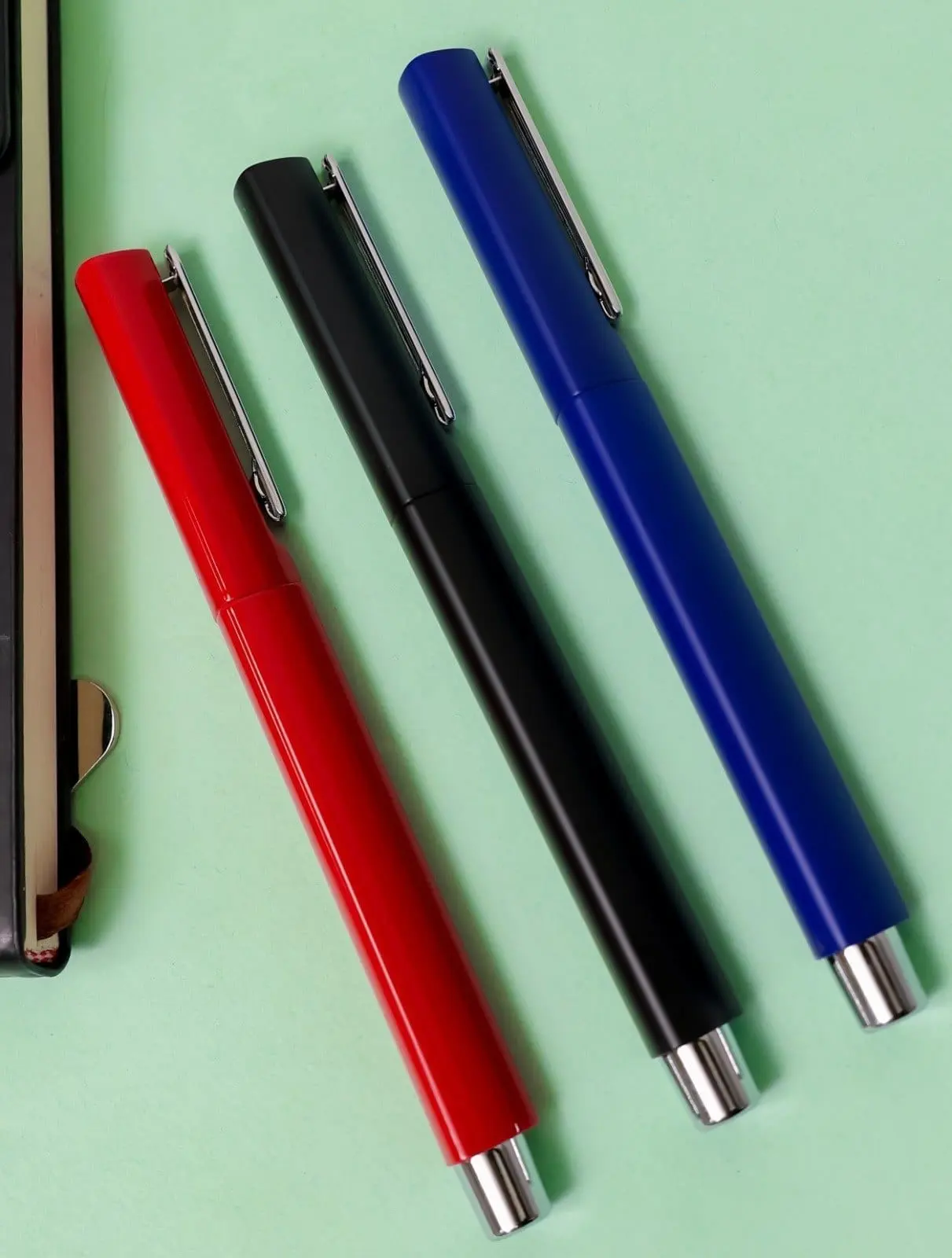 Red, black and blue metal pens with chrome clips and chrome ends on a light green surface, Hensa