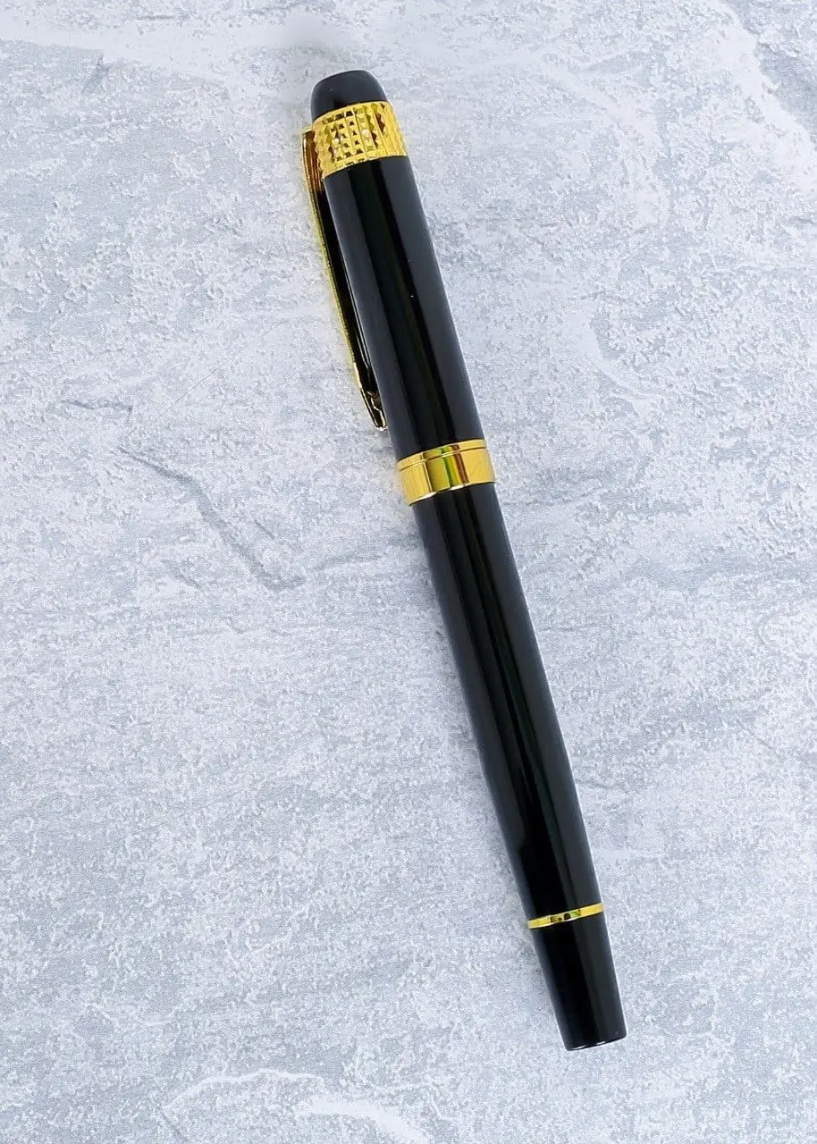 Glossy black metal pen with a gold clip and gold bands lying on a grey stone surface, Hensa