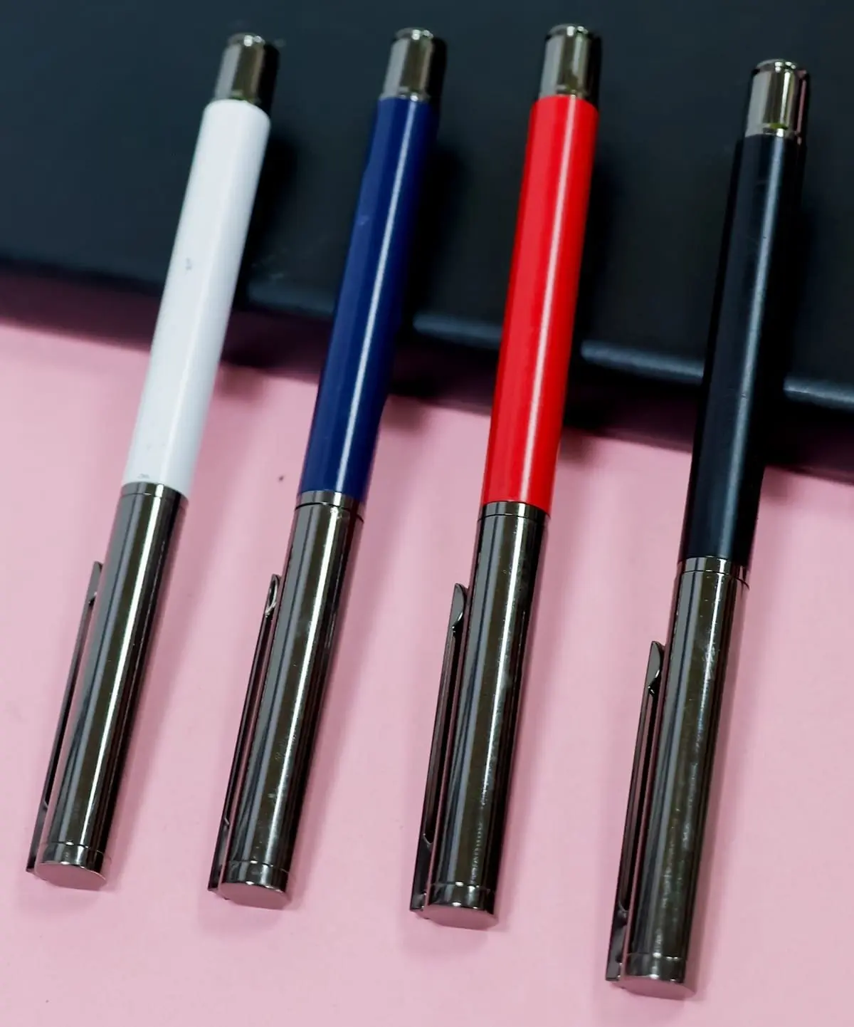 Four metal pens with gunmetal caps and white, navy, red and black barrels on a pink surface, Hensa
